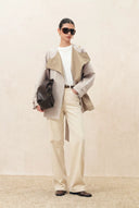 Woman wearing a taupe cropped trench coat with wide lapels, cream trousers and a dark shoulder bag