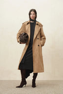Woman wearing a tan trench coat over a black asymmetric knit dress with brown shoulder bag and heeled boots